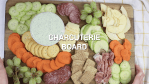 A wooden cutting board filled with crackers, cheese, salami, carrots, cucumbers, apples, and pears. Served with vegetable dip.
